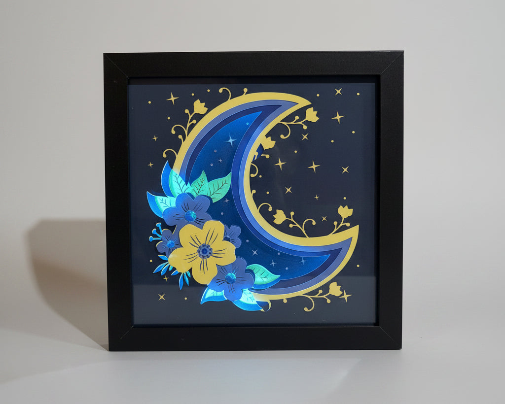Decorative wall art with a blue and yellow crescent moon and floral design in a black frame.