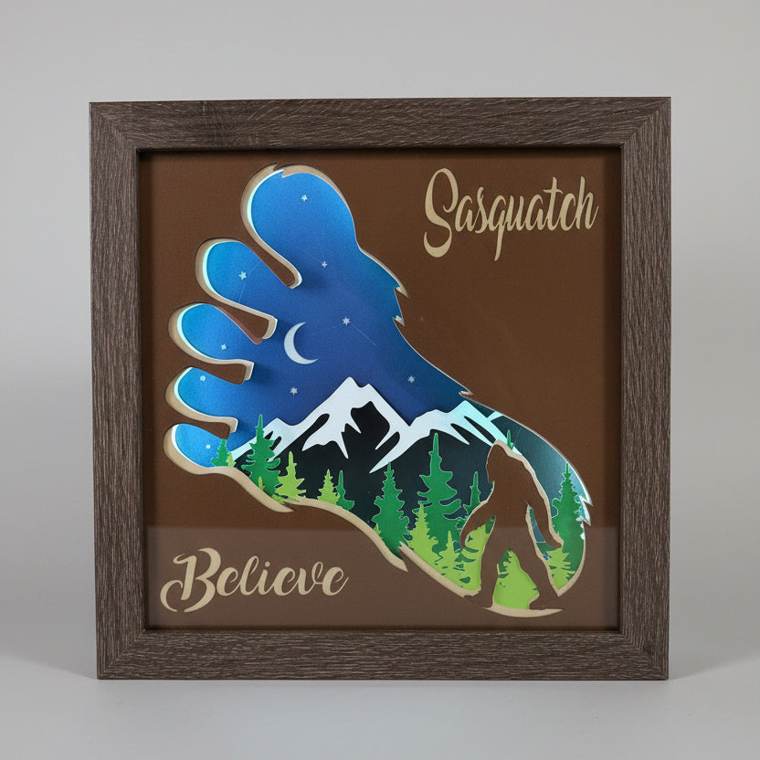 Framed artwork with a sasquatch silhouette, trees, and the word 'Believe' on a brown background.
